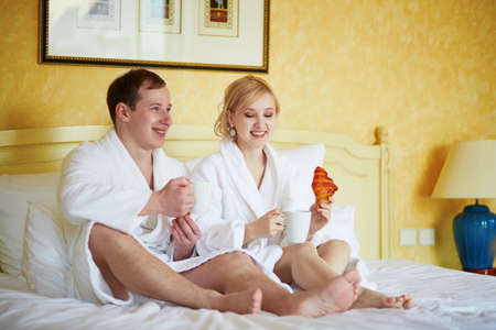 Happy young couple in white bathrobes drinking coffee together in bed at morning. Hotel, travel, relationships, and happiness conceptの写真素材