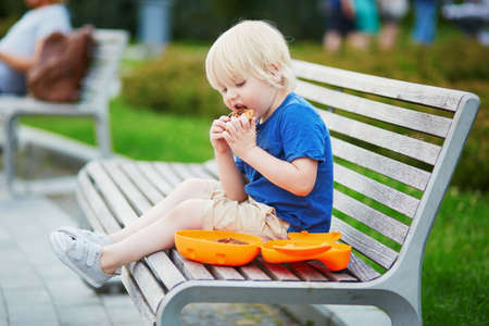 Adorable little boy sitting on the bench with lunchbox in park. Healthy food and snacks for kids. Child eating his breakfast or lunch outdoorsの写真素材
