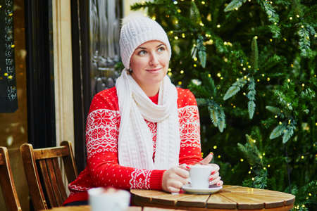Cheerful girl in red winter sweater drinking coffee in Parisian outdoor cafe decorated for Christmas