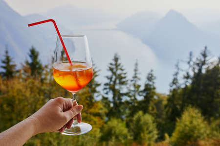 Woman holding glass with cocktail aperol spritz with scenic view to the lake Lugano from Monte Bre in Lugano, canton of Ticino, Switzerlandの写真素材