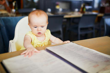 Little girl in restaurant. Baby sitting in high chair in cafe and reading menu. Eating out with kidsの写真素材