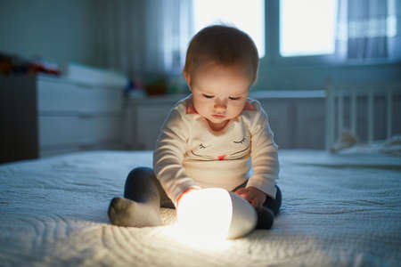 Adorable baby girl playing with bedside lamp in nursery. Happy kid sitting on bed with nightlight. Little child at home in the evening before sleepの写真素材