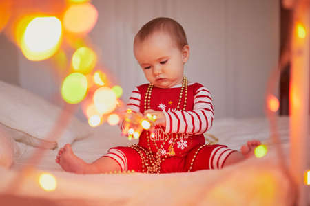 Happy little baby girl wearing pyjamas playing with light garland on her very first Christmas....
