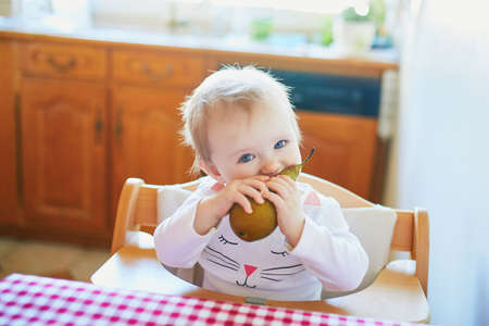 Cute baby girl eating pear in the kitchen. Little kid tasting solids at home. Baby led weaningの写真素材