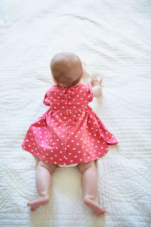 Six months old baby girl in pink dress trying to crawl. Little child in bright clothes lying on bed in nursery. Fashion for infantsの写真素材