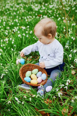 Cute little one year old girl playing egg hunt on Easter. Toddler sitting on the grass with many...