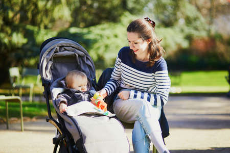 Young woman with toddler girl in pushchair walking together in Luxembourg garden of Paris, France. Mother and daughter in park on a sunny day. Student babysitter taking care of little kid