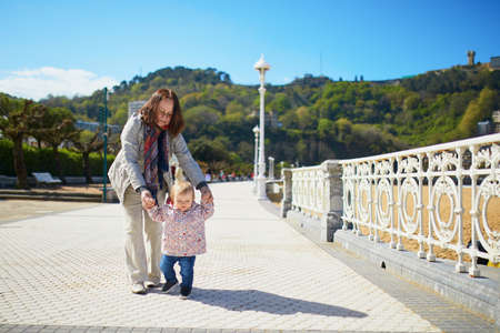 Grandmother helping granddaughter make her firts steps. Middle aged woman with little girl on the sea embankment. Toddler learning how to walkの写真素材