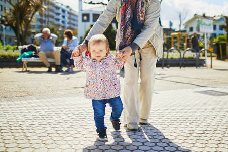 Grandmother helping granddaughter make her firts steps. Middle aged woman with little girl on the sea embankment. Toddler learning how to walkの写真素材
