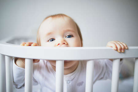 Adorable baby girl in co-sleeper crib attached to parents bed. Little kid sitting in cot