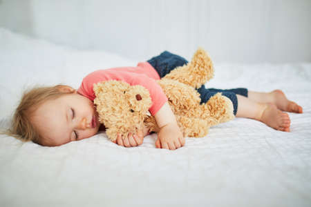 Adorable baby girl sleeping with her favorite toy. Little child having a day nap and hugging a teddy bear. Infant kid resting in nurseryの写真素材