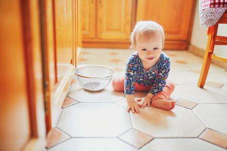Baby girl sitting on the floor in the kitchen and playing with kitchenware. Little child at homeの写真素材