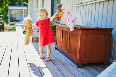 Adorable baby girl in red dress playing with doll and teddy bear outdoors on a sunny summer day. Little child having fun. Kid playing stuffed toysの写真素材