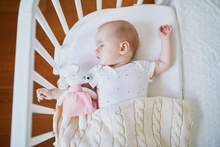 Adorable baby girl sleeping in co-sleeper crib attached to parents' bed with stuffed toy. Little child having a day nap in cot. Sleep training concept. Infant kid in sunny nurseryの写真素材