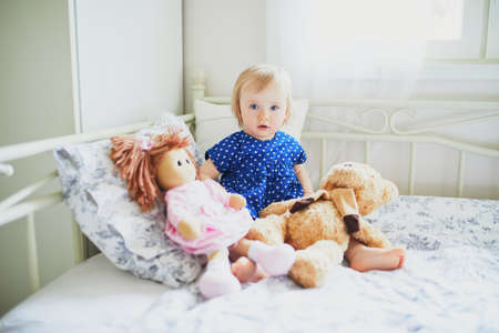Adorable baby girl in blue dress sitting on bed and playing with doll and teddy bear. Little child having fun in bedroom. Kid playing stuffed toysの写真素材