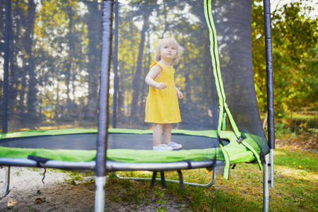 Adorable toddler girl in yellow dress having fun on a trampoline on a summer day. Little kid in bouncy house. Outdoor activities for kidsの写真素材