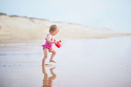 Adorable toddler girl playing with red watering can on the ocean beach. Outdoor activities for kidsの写真素材