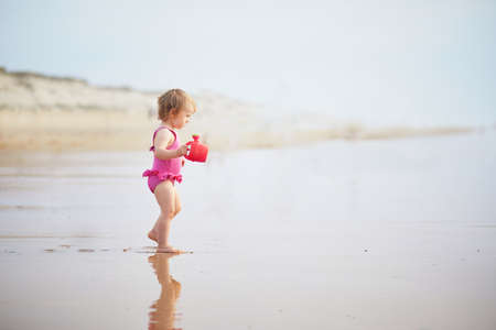 Adorable toddler girl playing with red watering can on the ocean beach. Outdoor activities for kidsの写真素材