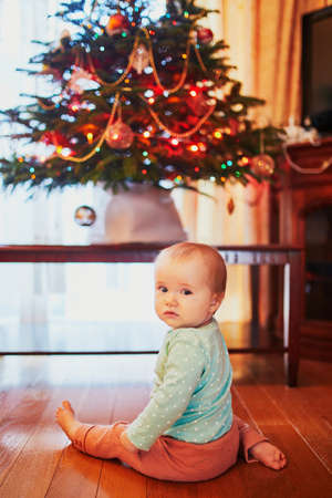 Baby girl sitting on the floor under Christmas tree. Little child crawling at home and exploring...