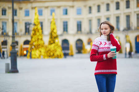 Happy young girl in warm red knitted holiday sweater walking with hot drink to go on a street of Paris decorated for Christmasの写真素材