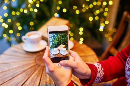 Woman taking photo of her coffee cup and Christmas present in cafe decorated for seasonal holidays