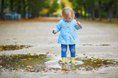 Child wearing yellow rain boots and jumping in puddle on a fall day. Adorable toddler girl having fun with water and mud in park on a rainy day. Outdoor autumn activities for kidsの写真素材