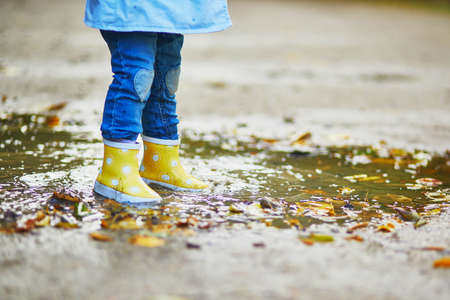 Child wearing yellow rain boots and jumping in puddle on a fall day. Toddler girl having fun with water and mud in park on a rainy day. Outdoor autumn activities for kidsの写真素材