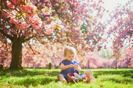 Cute little one year old girl playing egg hunt on Easter. Toddler sitting on the grass under cherry...