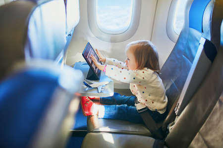 Adorable little toddler girl traveling by plane. Small child sitting by aircraft window and using a digital tablet during the flight. Traveling abroad with kids. Unaccompanied minor conceptの写真素材