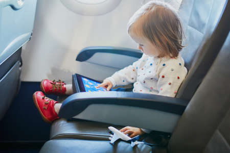 Adorable little toddler girl traveling by plane. Small child sitting by aircraft window and using a...