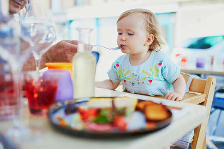 Adorable toddler girl sitting in high chair in cafe or restaurant. Father feeding his little daughter. Going out with kids conceptの写真素材