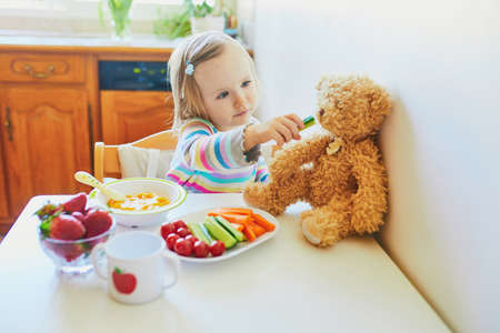 Adorable toddler girl eating fresh fruits and vegetables for lunch. Child feeding doll and teddy bear in the kitchen. Delicious healthy food for kidsの写真素材