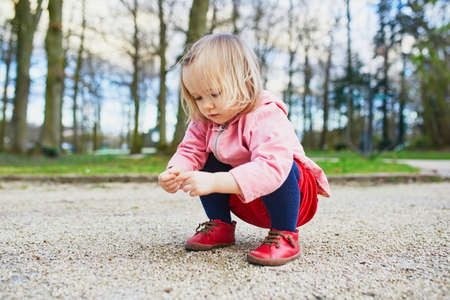 Adorable baby girl in park, sitting on hunkers and playing with little stones. Little child having...
