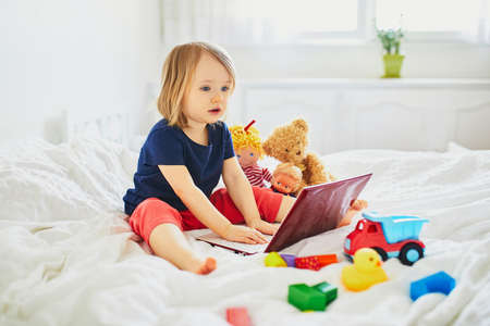 Toddler girl with laptop and toys in bed. Kid using gadget to communicate with friends or kindergartners. Education, distance learning or work from home with kids. Stay at home entertainmentの写真素材