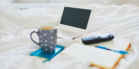 Laptop, cup of coffee, notebook and phone in bed on clean white linens. Freelance, distance learning or work from home conceptの写真素材