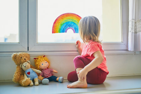 Adorable toddler girl attaching rainbow drawing to window glass as sign of hope. Creative games for kids staying at home during lockdown. Self isolation and coronavirus quarantine conceptの写真素材