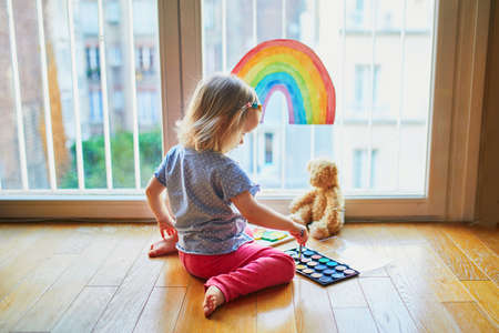 Adorable toddler girl painting rainbow on the window glass as sign of hope. Creative games for kids staying at home during lockdown. Self isolation and coronavirus quarantine conceptの写真素材