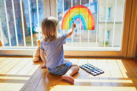 Adorable toddler girl painting rainbow on the window glass as sign of hope. Creative games for kids staying at home during lockdown. Self isolation and coronavirus quarantine conceptの写真素材