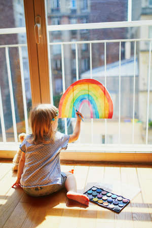 Adorable toddler girl painting rainbow on the window glass as sign of hope. Creative games for kids staying at home during lockdown. Self isolation and coronavirus quarantine conceptの写真素材