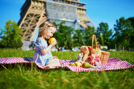 Cheerful toddler girl having picnic near the Eiffel tower in Paris, France. Happy child playing with toys in park on a summer day. Kid enjoying healthy snacks outdoorsの写真素材