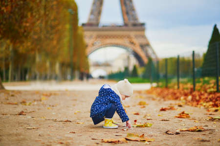 Adorable toddler girl in blue raincoat gathering yellow fallen leaves near the Eiffel tower in Paris, France. Happy child enjoying warm and sunny fall dayの写真素材