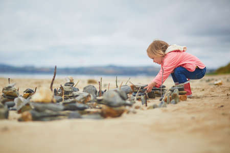 Adorable toddler girl playing with stones and building a town on the sand beach at Atlantic coast of...