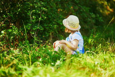 Adorable toddler girl having fun outdoors on a sunny summer day. Happy child sitting in green grass in the forest. Outdoor activities for kidsの写真素材
