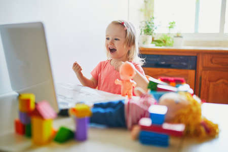 Happy toddler girl with laptop and toys. Kid using computer to communicate with friends, elderly relatives or kindergartners. Education or online communication for kids. Stay at home entertainmentの写真素材