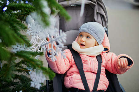 Adorable toddler girl sitting in pushchair near Christmas tree decorated with white baubles and snowflakes. Celebrating seasonal holidays with kidsの写真素材