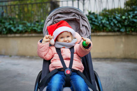 Adorable toddler girl sitting in pushchair in red Santa hat on a winter day. Celebrating Christmas and seasonal holidays with kidsの写真素材