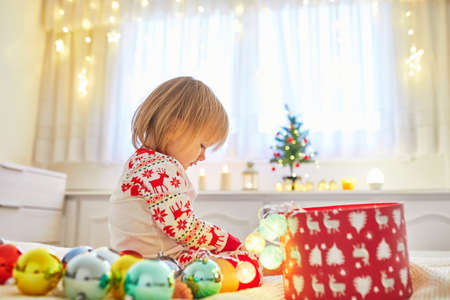 Happy little toddler girl wearing pajamas and playing with Christmas decorations on bed. Celebrating seasonal holidays with kids at homeの写真素材