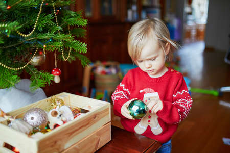 Adorable little toddler girl wearing holiday sweater decorating Christmas tree. Celebrating seasonal holidays with kids at homeの写真素材