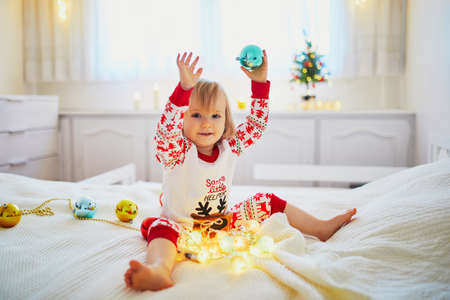 Happy little toddler girl wearing pajamas and playing with Christmas decorations on bed. Celebrating seasonal holidays with kids at homeの写真素材