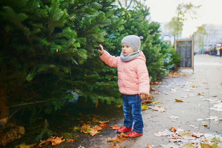 Adorable toddler girl selecting Christmas tree in outdoor shop or on market in Paris, France. Celebrating seasonal holidays with kidsの写真素材